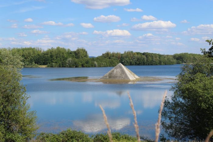 Etang entouré de verdure. On observe une pyramide en béton au milieu de l'eau.